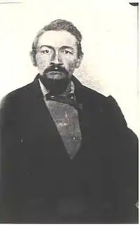Portrait of Ash Upson, seated wearing a dark suit, sweater and bow tie