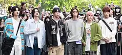 Six men standing in a row outdoors, dressed in colorful casual outfits, wearing a mix of oversized sweaters, cardigans, and jackets in pastel and neutral tones, with some accessorizing with glasses, crossbody bags, and layered clothing.