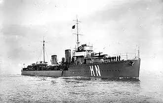 A black-and-white photograph of a naval destroyer at sea, viewed from the front-left side. The ship has two tall funnels, a central mast with rigging, and several sailors standing on the forward deck. Large white letters "K N" are painted prominently on the bow. The sea is calm and the sky overcast.