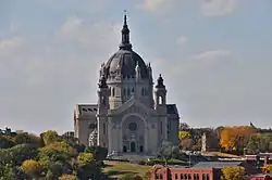 A towering gray stone cathedral with a round copper roof stands above autumn trees.