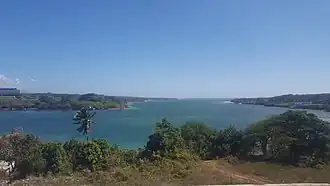 A photo taken from on top of a hill or cliff, overlooking a large bay. An entrance to the bay can be seen between flat landmasses on the right and left.