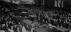 Black-and-white photograph showing a wide and busy street in a city center, with the outer lanes of traffic as well as the sidewalks filled by a massive crowd of people standing in celebration. Only the center two lanes of the road are being used by automobiles. In these center-two lanes is bumper-to-bumper automobile traffic, with the traffic consisting solely of dark-colored personal automobiles of an 1920s-era design. The vehicles are traveling bi-directionally in the North American-style norm of having vehicles drive on the righthand side of their respective direction