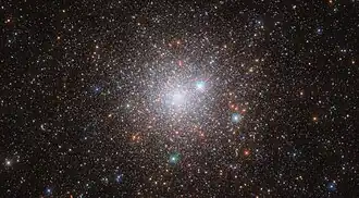 A cluster of stars in space. It’s bright in the centre, where the stars are densely packed together in the cluster’s core, and grows dimmer and more diffuse out to the edges, as the stars give way to the dark background of space. A few orange stars are spread across the cluster, but most are pale, bluish-white points of light. Three large stars with cross-shaped spikes around them lie between us and the cluster.