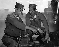 "Two men in military uniform at an Army base, ponder over instructions on how to change parts of a military truck."