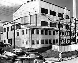 A large four-story building. The chimney is in the background. There are power poles and power lines in front.