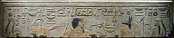Light gray stone surface with carved and painted images of two woman, a falcon-headed god, a black-haired man with a long goatee, a jackal-headed god, and Egyptian hieroglyphs inscribed along the top