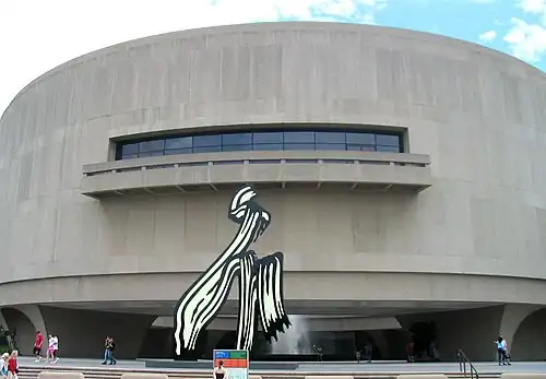 A large, convexly curving, 3 story tall, gray wall punctured on the second floor by a long strip of windows and a short balcony. A large black and white sculpture of a paint stroke sits in front of the building, stretching from the ground to the balcony