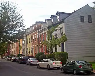 A row of attached houses along a street, seen from further down along the opposite side, on the right of the image. All are of brick, in various colors, three stories high, with gabled roofs and gabled dormer windows. There are tall trees on the right and cars parked along the same side of the street as the houses.