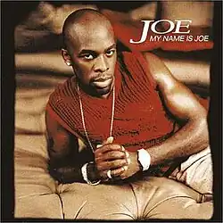 An image of a man in a red tank top, sporting a platinum necklace, watch, and rings, lying down on a beige bed.