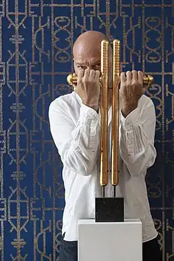 Kendell Geers, South African-Belgian artist, standing in front of patterned artwork while holding a sculptural piece.