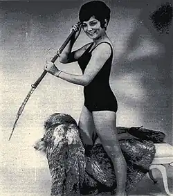 Photograph of a model in a bathing suit standing but with one knee resting on a fur covered bench and holding a harpoon.