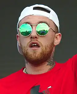 A tattooed Caucasian man holding a microphone smiles while rapping.