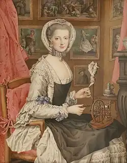 A young woman in a simple white dress with a black apron and a white cap is sitting before a background of small paintings. She is weaving on a small loom.