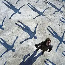 The album cover depicts a man holding a gas mask looking above as he stands among the shadows of floating figures.