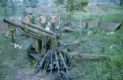 A damaged artillery piece sits in the foreground with rifles resting against it. In the background white soldiers stand next to a hootchie.
