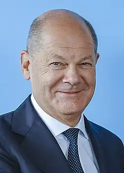 An older man partially smiling with short grey hair, clean shaven, and a blue suit