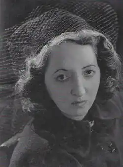 A young woman looks into the camera with a serious expression, her short, wavy hair topped by a hat with veiling