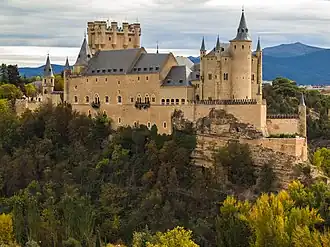 A castle high on a rocky peninsula above a plain. It is dominated by a tall rectangular tower rising above a main building with steep slate roof. The walls are pink, and covered with a sculptural pattern. There is a variety of turrets and details.