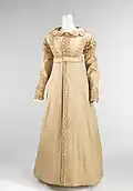 A headless mannequin wearing a light beige floor-length coat that buttons up the middle with three lines of decorative, zigzagging cording along the fitted bodice, long sleeves, and scalloped collar