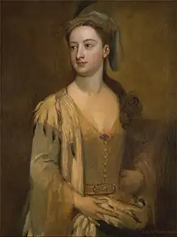 A woman sitting, looking to the left, wearing a golden dress and a floppy hat