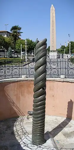Photograph of a bronze spiral column, with an obelisk in the background