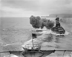 Two ships in line astern; the rear one with a prominent cloud of dirty-grey gunsmoke beside it