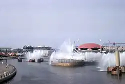 A lake with fountains and sculptures. Futuristic buildings line the far shore.