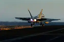 Photograph from behind a twin-engined jet fighter. The aircraft's wheels are on the surface, but the engines are still active, and a hook on the underside of the aircraft is in contact with the surface and trailing sparks