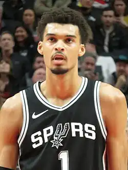 Wembanyama in 2022. He is a tall, black man with curly hair, a moustache and goatee, wearing a white Metropolitans 92 sports jersey.