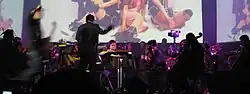 An orchestra performs a concert in a musical theatre, featuring music from the video game; in front of a screen showing people dressed as characters from the game.