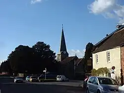Street scene with a church and spire central to the picture. To the right is a yellow building with a pub sign. To the left is a large tree with a signpost in front. Several cars.