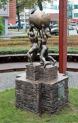 Four figures stand in a circle holding up the symbol of Wikipedia, which consists of a sphere made of jigsaw puzzle pieces, with few pieces missing, symbolizing that the work of the encyclopedia will never be finished