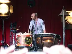 Champion resting his hands in a timpani, wearing a light blue shirt and purple scarf